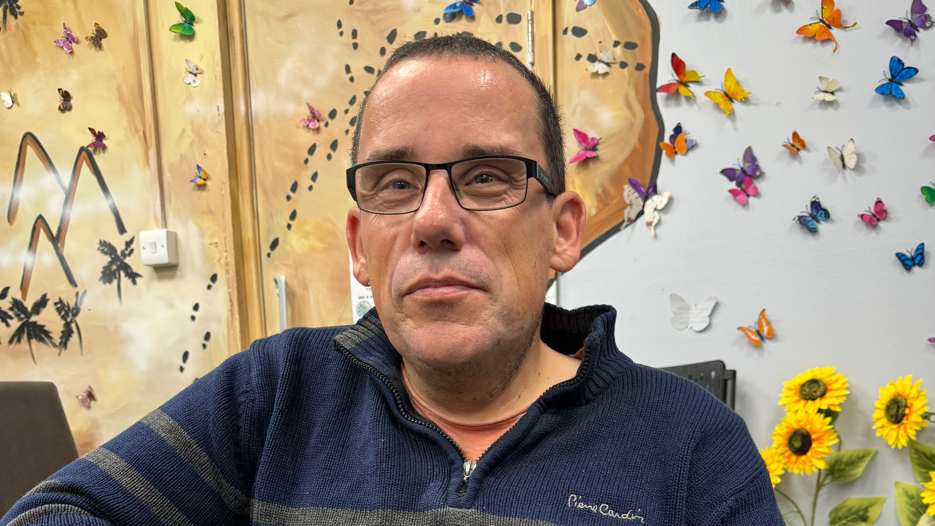 John Smith wearing glasses and a blue and green jumper. There are flowers and butterflies on a wall behind him. 