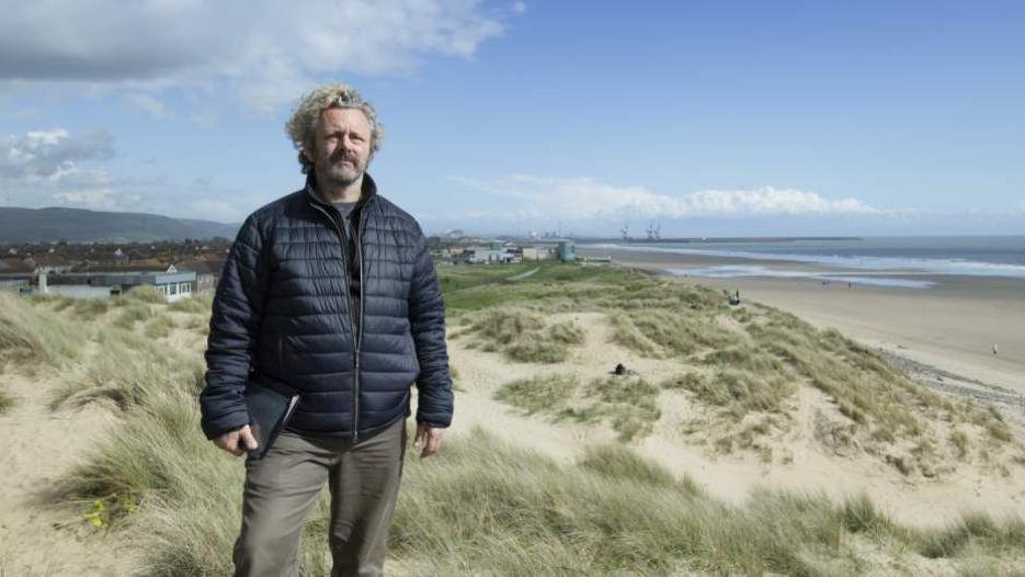 The actor Michael Sheen stands on some sand dunes looking into camera wearing a navy padded jacket and holding a folder.