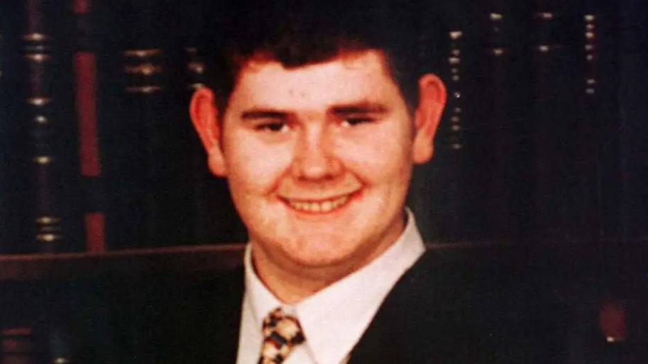 Young man wearing a suit and tie standing in front of a bookcase smiling at the camera