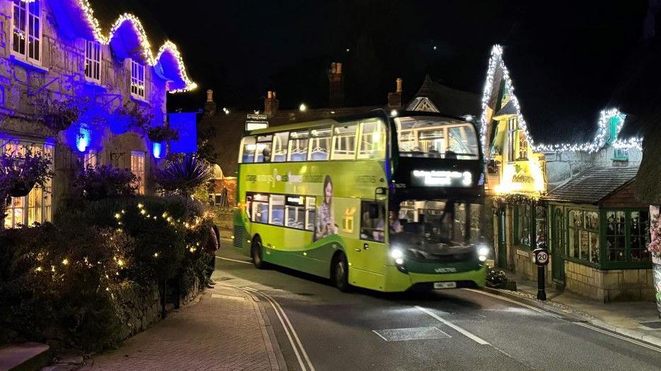 Green bus travelling through road with buildings decorated in lights either side - it is night.
