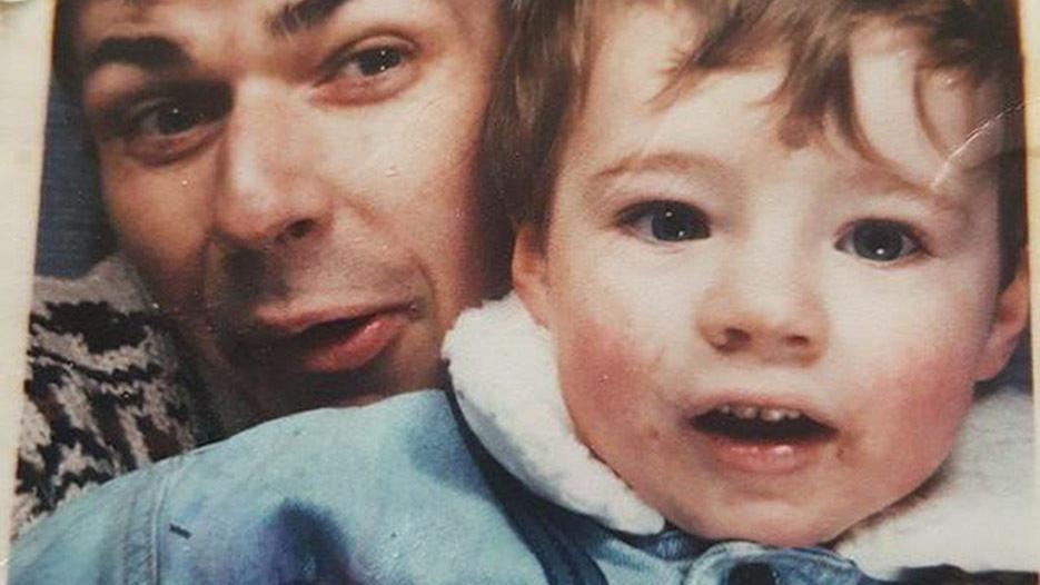 Mr Evans making a silly face with baby Jason. His father is on the left and is clean shaven  with dark hair and wearing a grey jumper while Jason who looks to be near toddler-age, wears a blue denim jacket with a white fluffy collar.