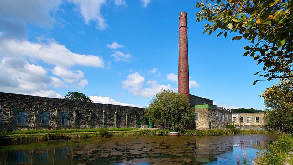 Queen Street Mill's chimney against a blue sky with water to the front and a large stone wall to the left 