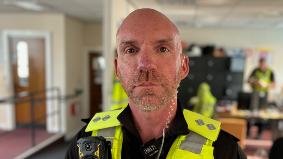 A police officer with a small white beard wearing a high visibility jacket and body camera in an office