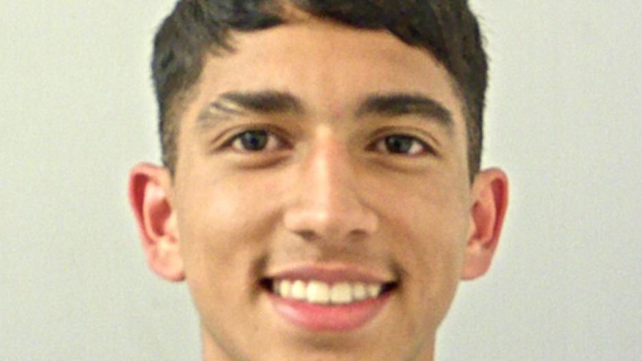 a closupe image of a young man with black hair, smiling. There is a neutral background.