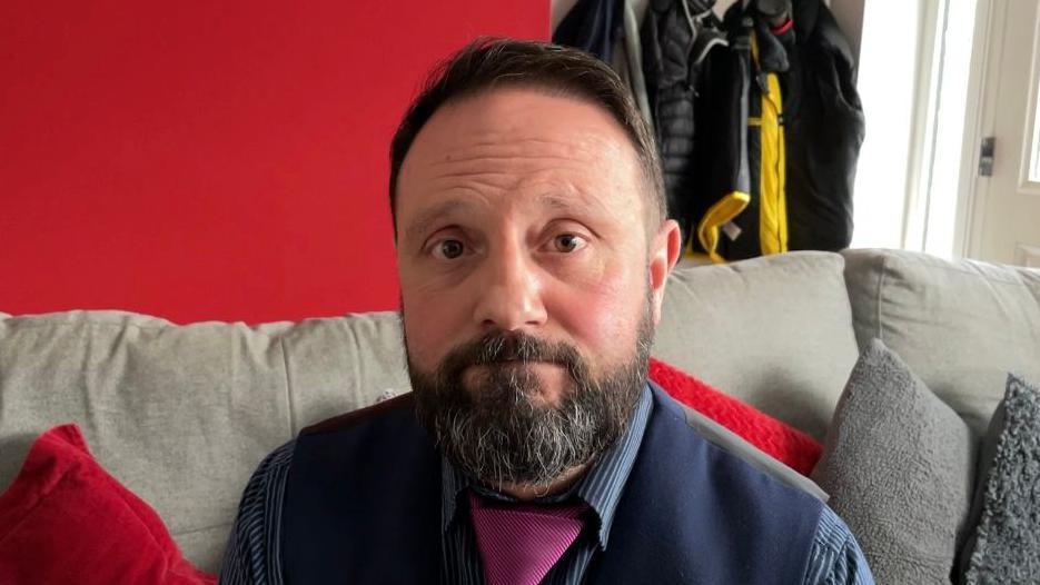 John Newcombe looks directly at the camera, he has dark brown hair and a beard and moustache, he is wearing a navy waistcoat with a blue pinstriped shirt and a pink tie. He is sitting on a grey sofa, the wall behind him is red.