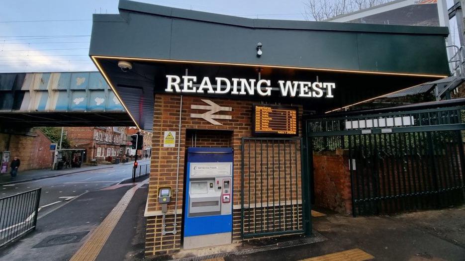 Outside of Reading West station. A lit-up sign reads 'Reading West' and there is a ticket machine beneath it.