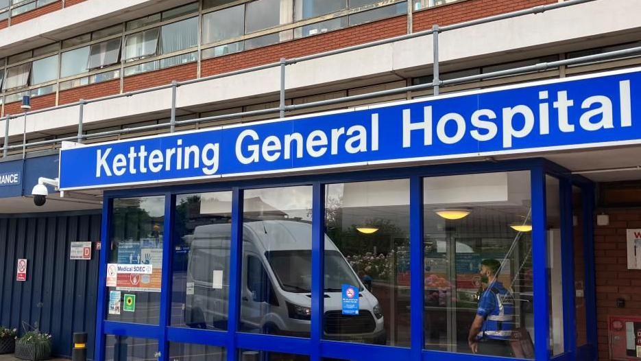 The blue framed entrance to Kettering General Hospital with a blue sign above the door. A security guard stands inside the entrance. 