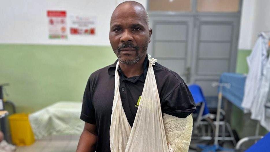 A man in a black t-shirt and jeans sits on a hospital bed. He wears a sling and a bandage around his arm.