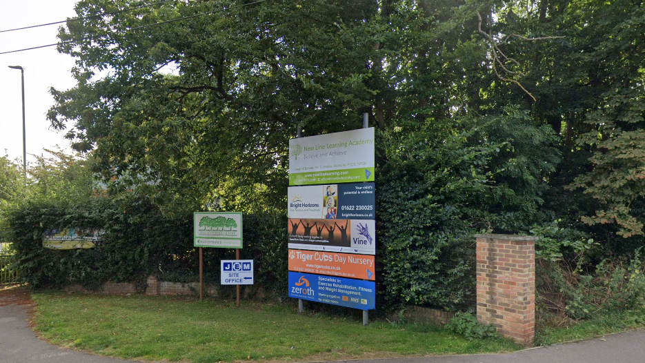 Signage on a grass verge advertising New Line Learning Academy, in green, ans a few other schools. The other signs are blue, orange, and navy and white.
