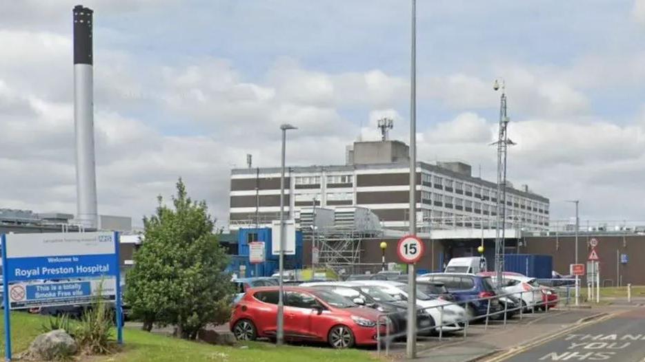 Sign which reads 'Welcome to Royal Preston Hospital' with a 15-mile speed limit sign on the road and parked vehicles in a car park in front of the hospital building.