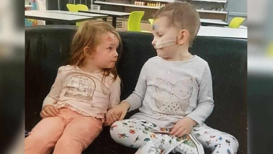 Two little girls are sat on a chair holding hands. They are looking directly at each other. They are both around three or four years-old and wearing leggings and a long sleeve top. 
The child on the left has brown hair, the child on the right has a feeding tube in her nose and very little hair. 