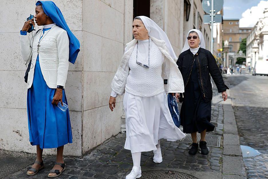 A Nigerian reverend sister wearing a blue habit on the phone as two other sisters wearing black and white habits walk by on the streets of Rome.