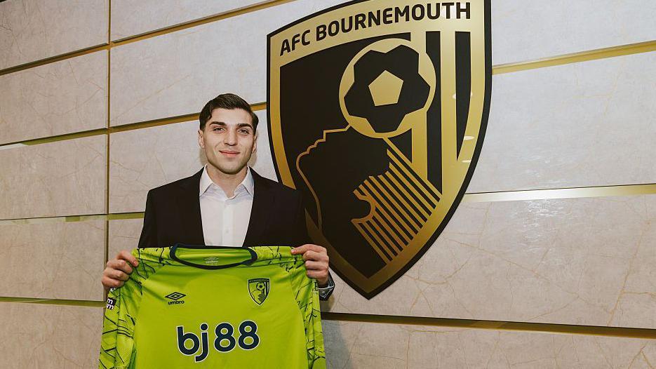 New Bournemouth signing Christos Mandas holds up a jersey by the club crest