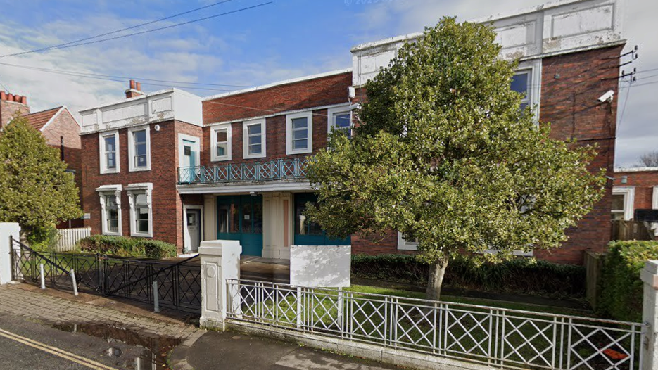 A two-storey brick building with a tree in front. There is a road in front with a puddle to the left of the image.