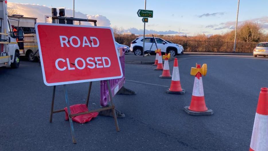 A 'road closed' sign and some traffic cones bordering off a road on a roundabout.  There is a white car and van in the background.