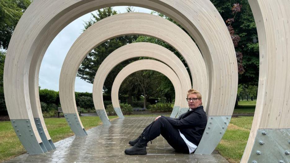 Sculptor Charlotte Holmes sits with her back against one of the sculpture's wooden arches, and looks left towards the camera. She has short, dyed blond hair, dark-rimmed glasses and is wearing a black jacket over a white top, with dark trousers and black boots.