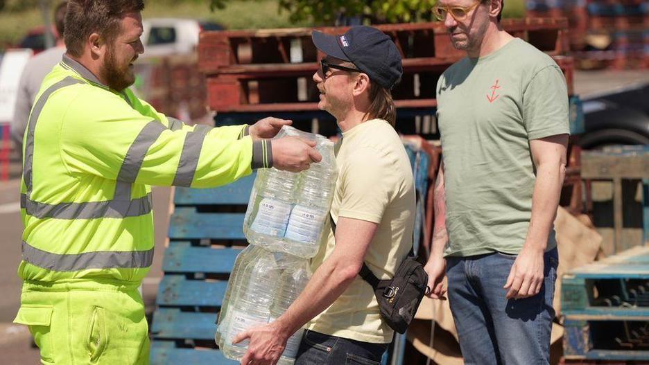 Two men in t-shirts are handed bottled water by a Southern Water worker, who is wearing a hi-vis jacket and trousers.