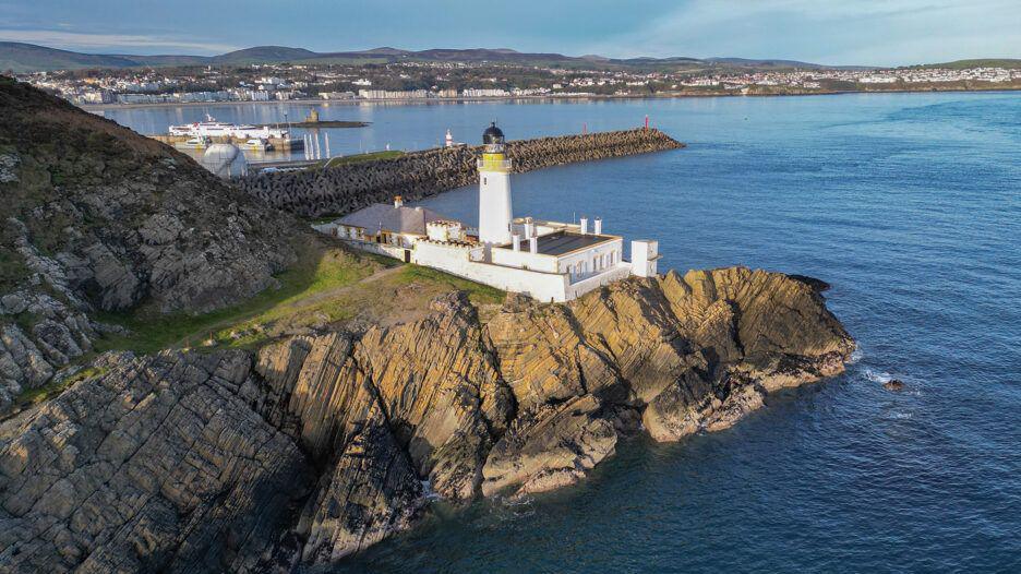 An aerial view of Douglas Bay, with a lighthouse in the foreground on rocks and the Douglas Promenade and Onchan Head in the distance.