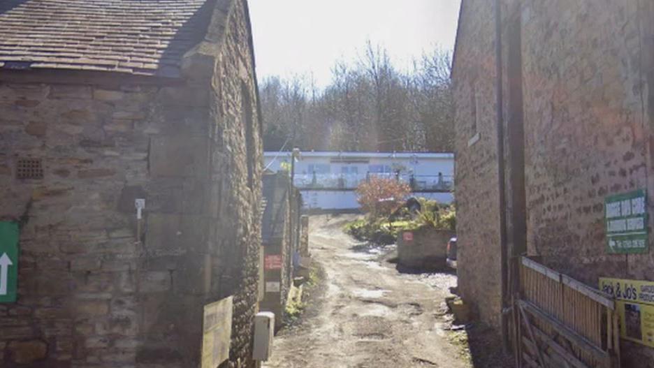 Through an alleyway between two brick buildings the cafe can be seen - it is a long while building with chairs outside on decking. 