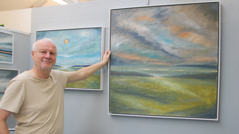 Tony, smiling to camera, and holding up a large painting of a landscape of green fields, and blue and orange sky