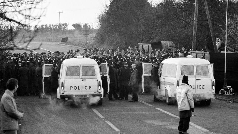 Black and white archive photograph of pickets and police clashing at Daw Mill Colliery, near Nuneaton, Warwickshire, on 27th March 1984. There are two police vans in the foreground and several rows of police officers.