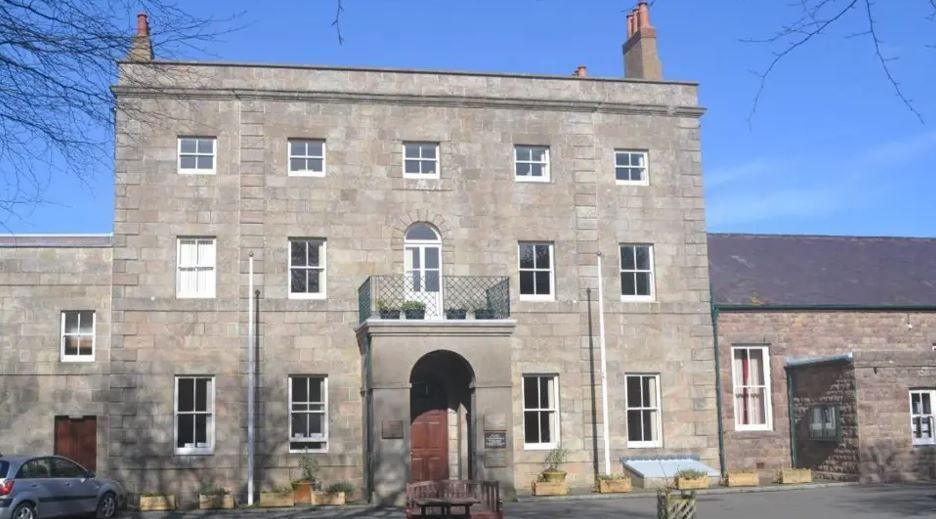 A three-storey grey building with windows on each floor and a brown door under a portico and a balcony on the first floor with a car parked to the left.