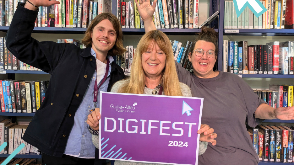 Three people stood in front of three bookshelves side by side. The person on the left is wearing a blue shirt with a white vest underneath and a blue jacket. The person in the middle is wearing a grey cardigan and is holding up a sign that says DIGIFEST 2024 in white writing on a purple background. The person on the right is wearing a grey t-shirt. All three people are smiling and the people on the left and right have their hands up in excitement. 