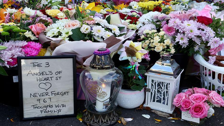 Flowers, candles and lantern at a makeshift memorial allowing people to pay their respects to the victims of the Southport knife attacks. There is also a framed poster which reads: The 3 Angels of Heart St Never Forget 9 6 7 Thoughts with Families.