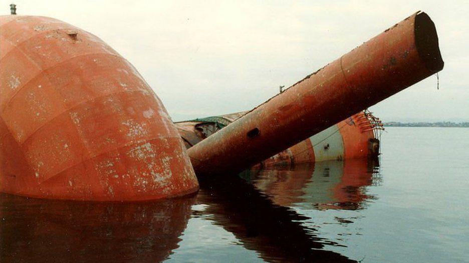 A giant red and white tube with a large round float on the bottom pokes up from a calm sea. Another tube is jutting out.