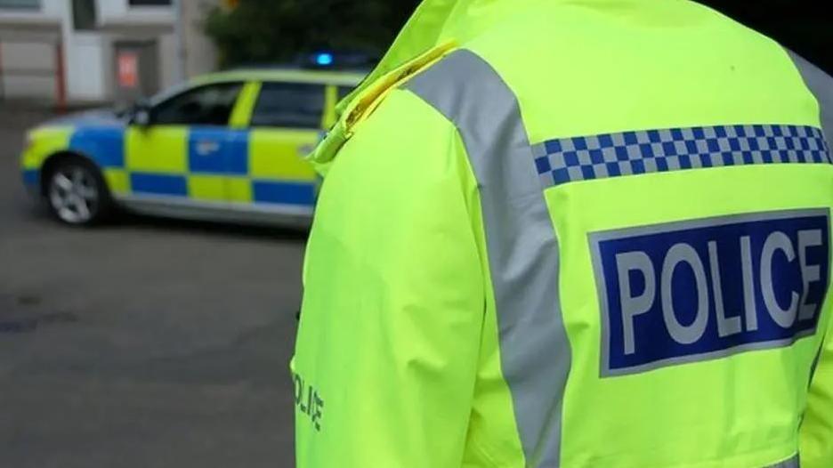 Police officer in hi-vis jacket taken from the back with a police car in the background.