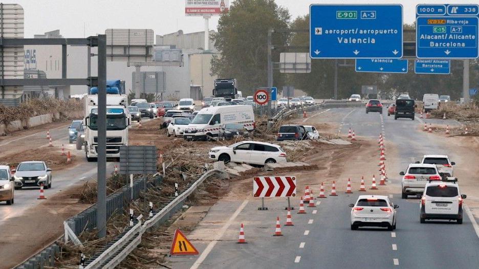 The A3 - a main road between Valencia and Madrid - is seen partially closed off, with some vehicles swept into mud and the central reservation. Some cars can pass on a section of road that is coned off in both directions. Above the road are motorway signs pointing to Valencia Airport and Barcelona