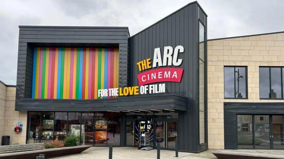 A black building frontage with The Arc Cinema logo on the side. There are a number of multi-coloured vertical panels on its left hand side.