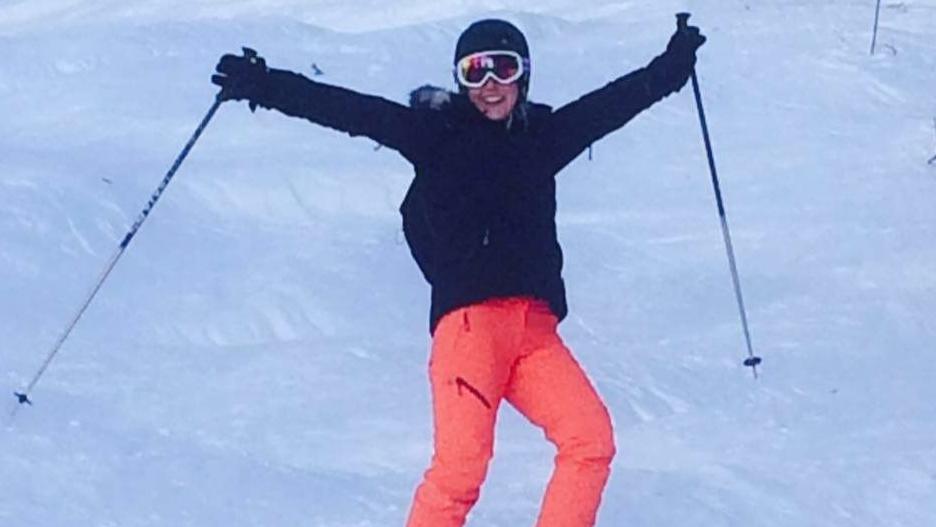 Becky Williams on snow in ski clothes and helmet, holding ski poles aloft