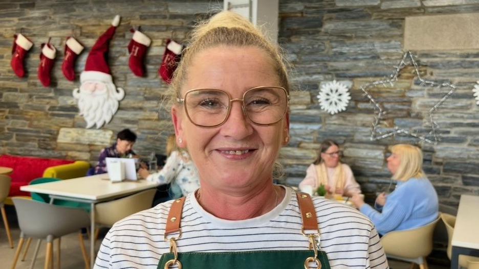 A lady wearing glasses smiles. She is standing in a cafe, a number of tables and cafe customers are in the background. She is wearing a white striped shirt, with an overall over it.