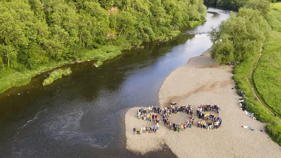 People form the letters SOS on the banks of the River Wye. There are trees either side of the river