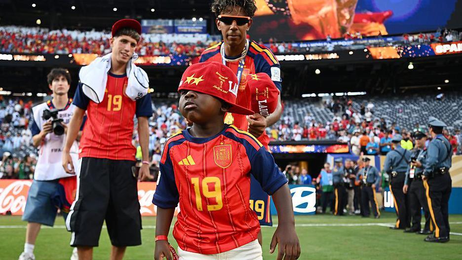 Kenye wears a number 19 shirt and a cap partially covering his eyes. His brother is behind him