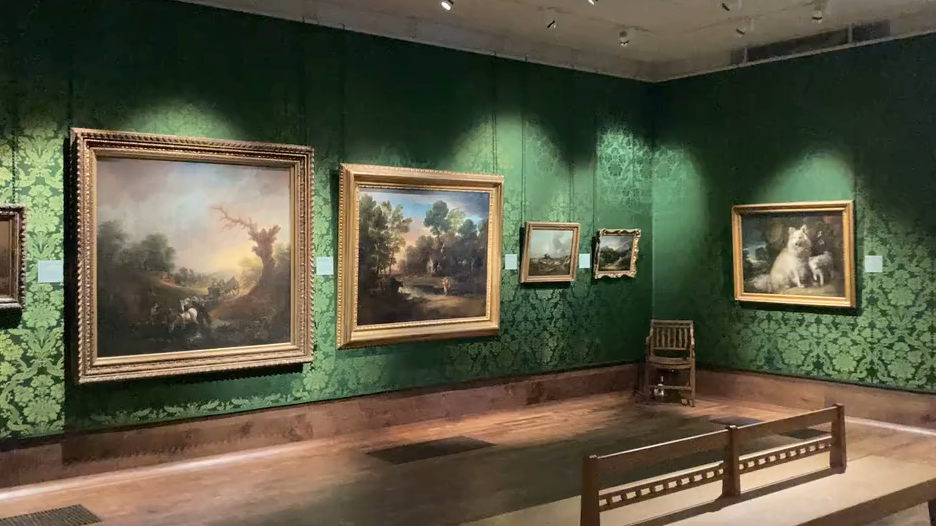 Large landscape paintings in gold frames in a darkened art gallery, decorated with dark green floral patterned wallpaper 