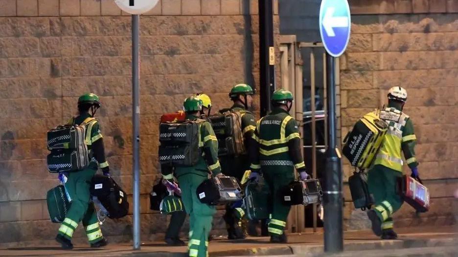 Ambulance personnel in green and yellow overalls and helmets, carrying bags. They are walking past a high stone wall and a one-way sign, responding to 999 calls from Manchester Arena.