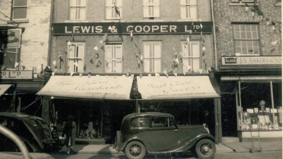 A black and white photograph of the double-fronted shop with bunting and awnings above the windows.  There is dark coloured car parked in front of the shop.