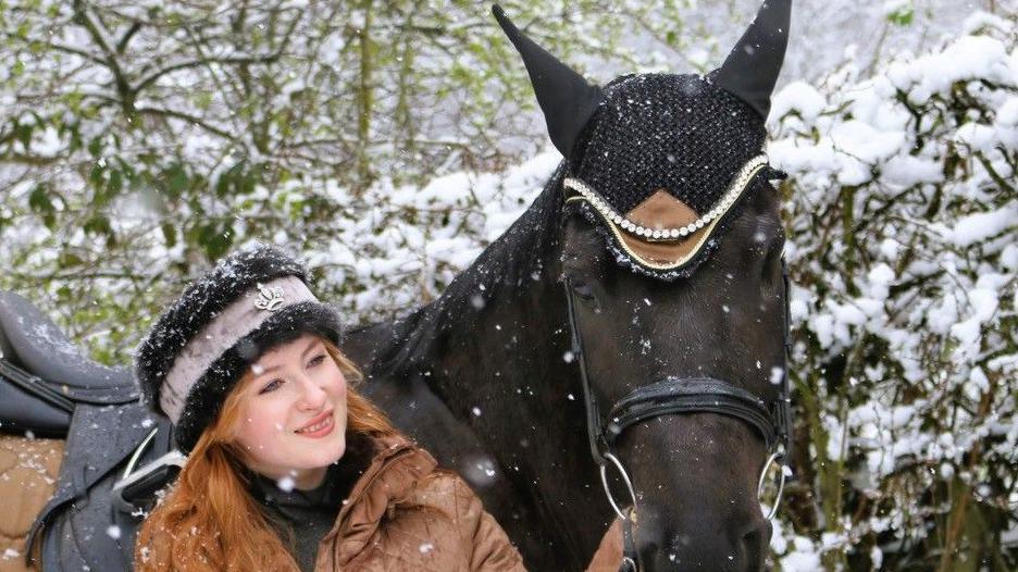 Alanna Clarke standing beside her horse Ella, in the snow.  Alanna wears furry hat and a tan padded jacket, she is wearing her auburn hair down under the hat, and holds the horse's bridle.  The horse wears an ornate headdress and in the background are green snow covered branches.