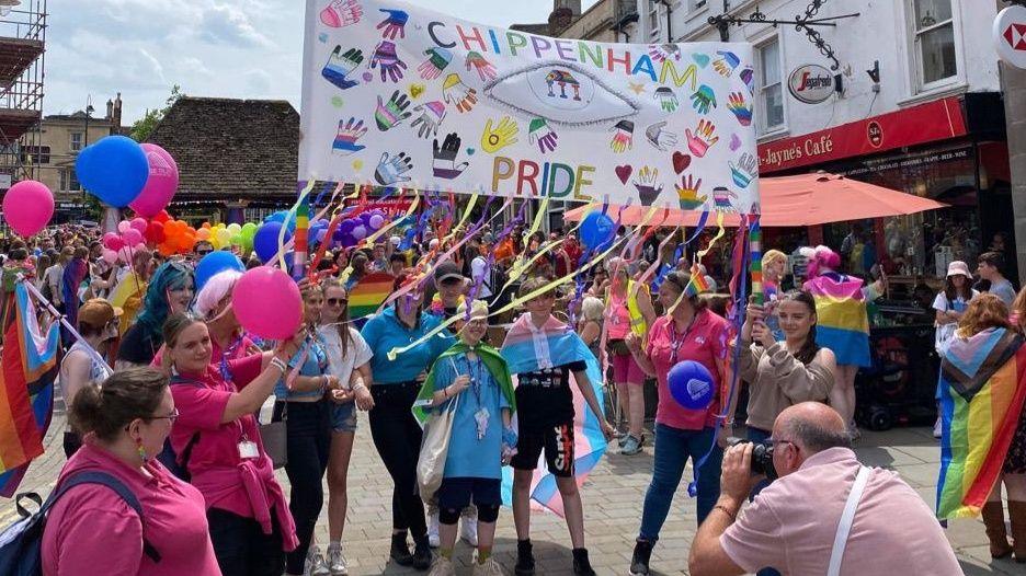 Chippenham Pride 2023, people surrounded by multi-coloured balloons and flags with a group in the centre of the picture carrying a pride banner