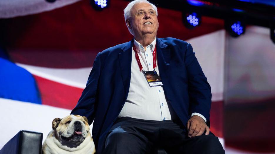Jim Justice sitting down beside a bulldog on a stage in front of an American flag