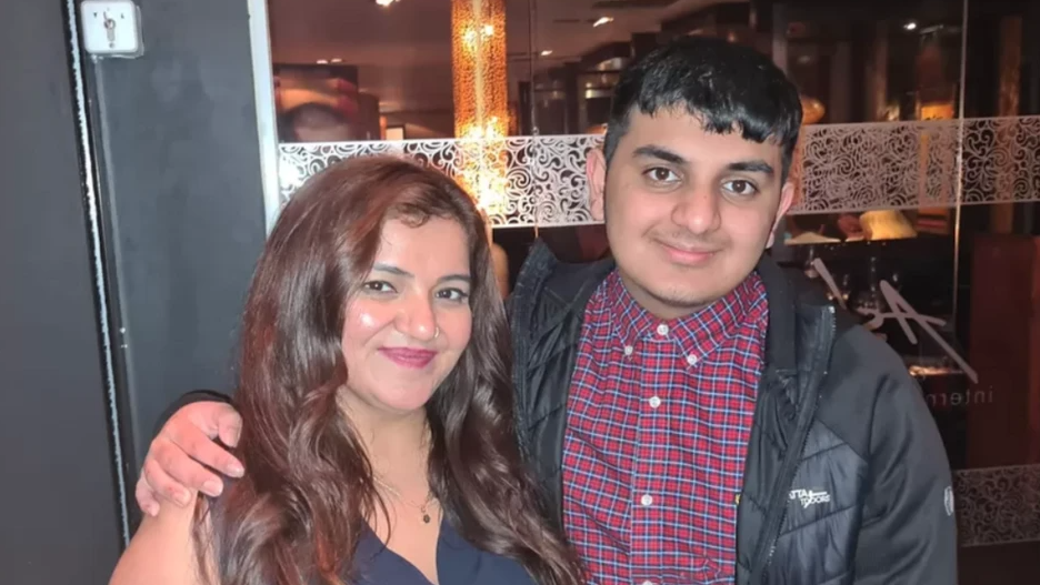 Pooja and Ronan are stood outside a restaurant. Ronan has his arm around his mother. He is wearing a red checked shirt and black jacket. She has long brown hair and is wearing a sleeveless top.