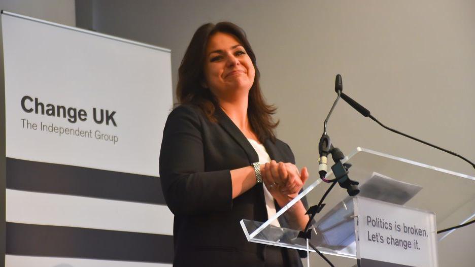 Heidi Allen, a woman with shoulder length black hair, dressed in a black suit, smiles and clasps her hands on a Change UK party podium. The slogan on the podium says "Politics is broken> Let's change it".