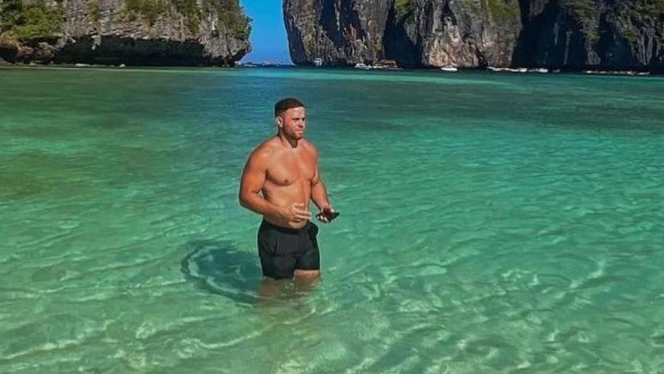 Corey Beavis stands in shallow water in the sea in Thailand. The water is a clear blue green and rocks rise up from the sea behind. He is wearing black swim shorts.