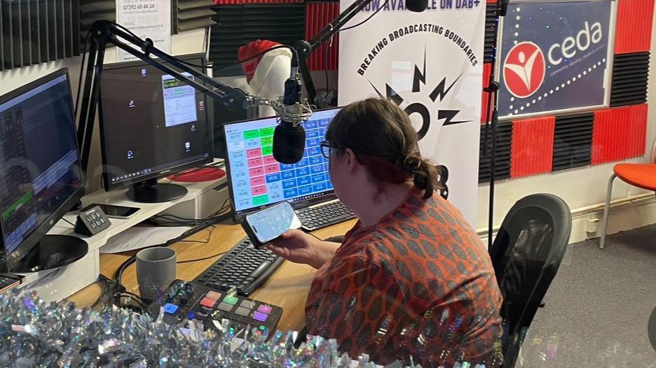 A female presenter sits in a radio studio reading out online messages from listeners. 