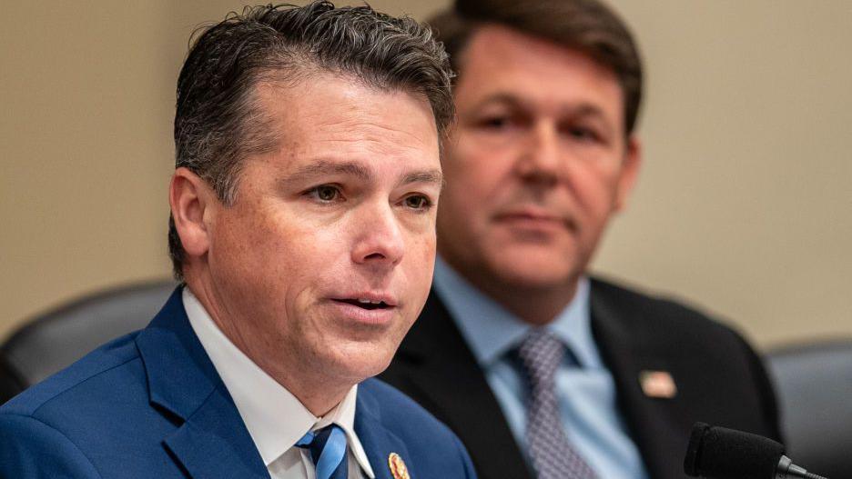 Headshot of Brendan Boyle wearing a suit and tie. Another man is sitting behind him out of focus.