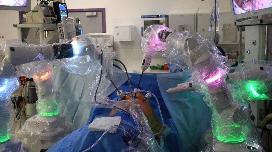 A person has been covered up by blue material while they are lay in an operating theatre. There are robotic arms, wrapped in plastic, around the person. Some of these arms look to be going into the person's chest. The machines have been lit in different colours like pink, blue and green.