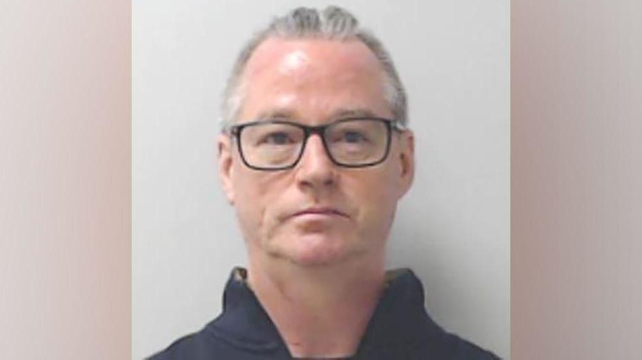 Man with short greying hair, wearing dark-rimmed glasses, looking at camera, wearing a dark top, in a police mugshot.