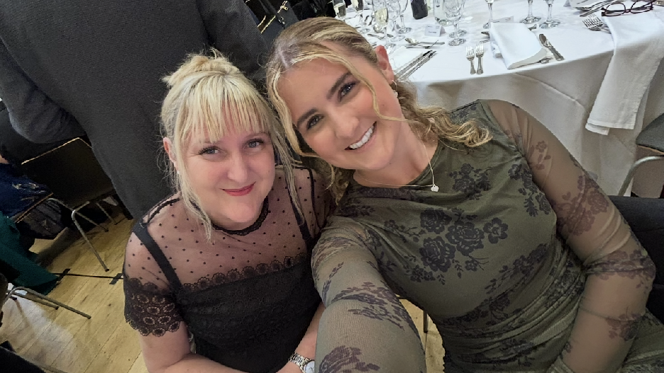 A selfie of me and my mother smiling at an awards ceremony in Manchester. My mother, Sian, is on the left and is wearing a black mesh dress. She has blonde hair. On the right, I am wearing a long sleeve green floral dress. I have light brown hair.